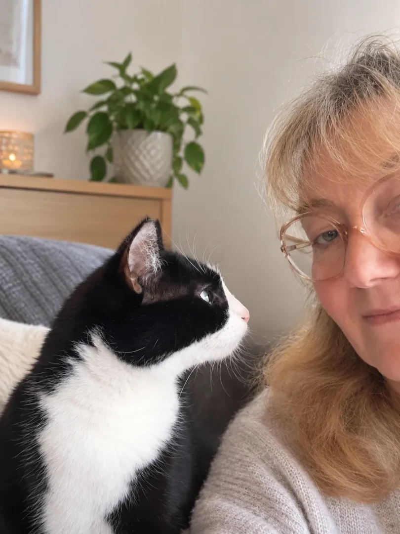 Chat noir et blanc observant de près une femme à lunettes, installés sur un canapé gris près d’une plante verte
