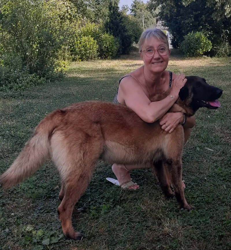 Femme souriante à lunettes accroupie dans un jardin, serrant dans ses bras un grand chien au pelage fauve
